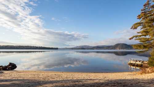 A serene lake scene with calm waters, sandy shore, and distant mountains under a clear blue sky.