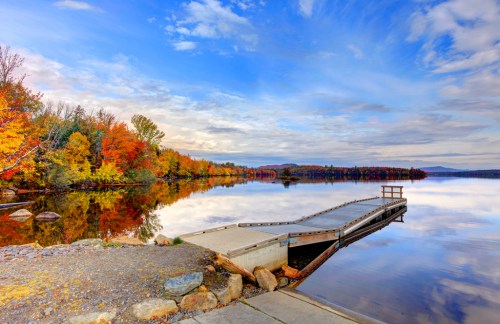 A serene lake scene with a wooden dock, surrounded by vibrant autumn foliage and a clear blue sky.