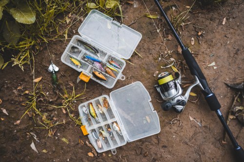A fishing setup with tackle boxes filled with lures and a fishing rod on the ground among grass and dirt.