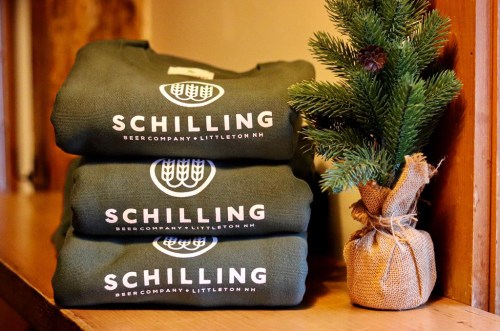 Three stacked green sweatshirts with "SCHILLING BEER COMPANY • LITTLETON NH" logo, next to a small pine tree in burlap.