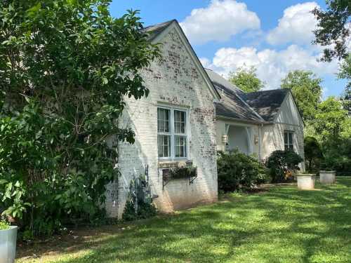 A charming white brick house surrounded by greenery and a well-maintained lawn under a blue sky with fluffy clouds.