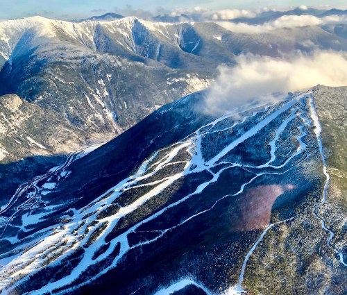 Aerial view of snow-covered mountains with winding ski trails and clouds in the background.