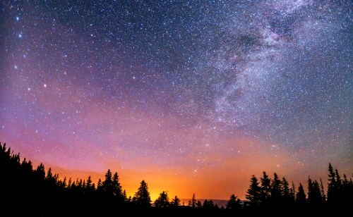 A starry night sky filled with vibrant colors, silhouetted trees in the foreground, and a glowing horizon.
