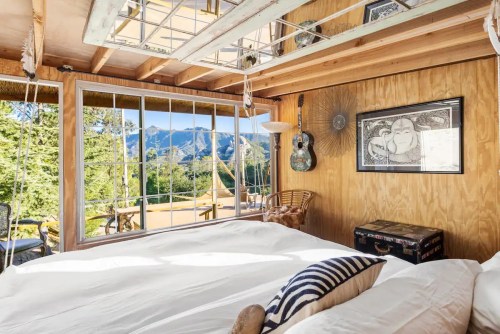 Cozy bedroom with a large window showcasing mountain views, featuring a bed, guitar, and wooden decor.