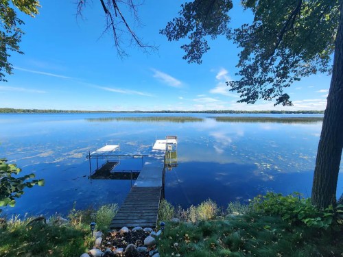 A serene lake view with a wooden dock, surrounded by trees and clear blue skies reflecting on the water's surface.
