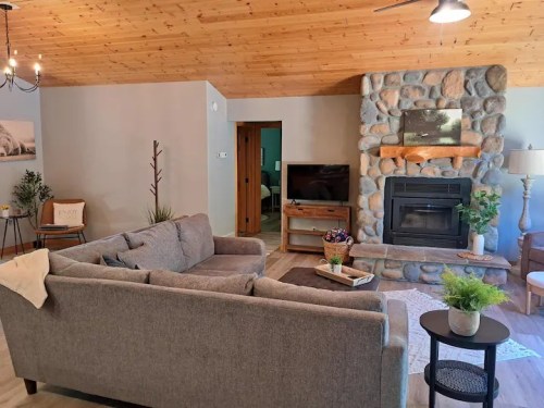 Cozy living room with a stone fireplace, gray sofa, wooden ceiling, and a TV, creating a warm, inviting atmosphere.
