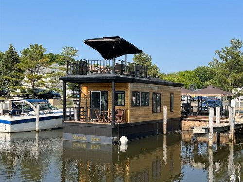 A modern houseboat named "Shoreside Haven" docked in a marina, featuring a rooftop deck and surrounded by trees.