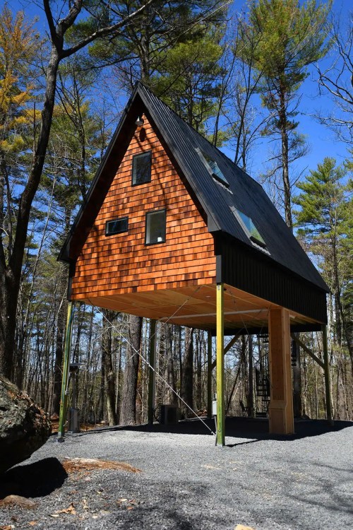 A modern A-frame cabin elevated on stilts, surrounded by trees and blue sky, with wooden shingles and large windows.