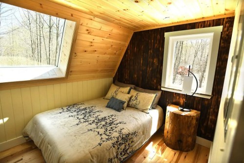 Cozy bedroom with a wooden ceiling, a bed with floral bedding, and a large window overlooking a forest.