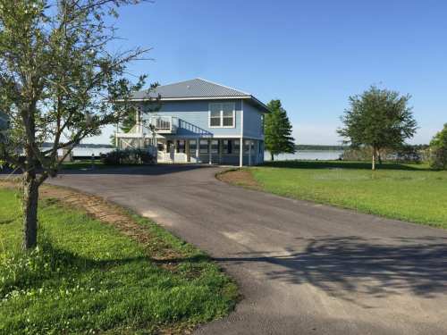 A two-story blue house by a lake, surrounded by green grass and trees, with a winding driveway leading up to it.