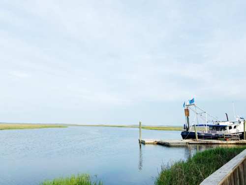 A calm waterway with a boat docked at a pier, surrounded by grassy marshland under a clear sky.
