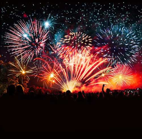 A vibrant display of colorful fireworks lighting up the night sky, with silhouettes of people watching below.