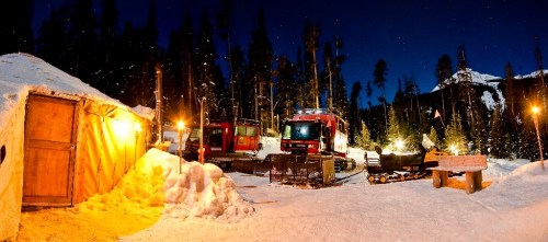 A snowy landscape at night featuring a wooden cabin, snowplows, and trees under a starry sky.