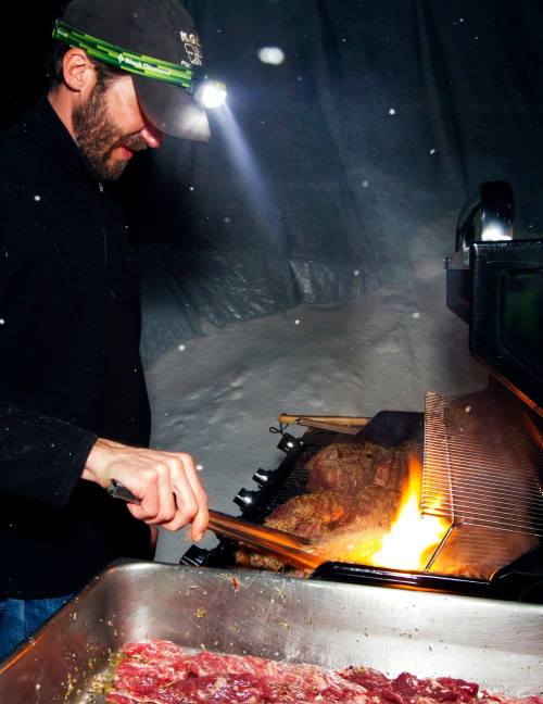 A person grilling meat outdoors in the snow, wearing a headlamp, with flames rising from the grill.