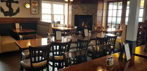 Cozy restaurant interior with wooden tables, chairs, and a fireplace, featuring nautical decor and large windows.