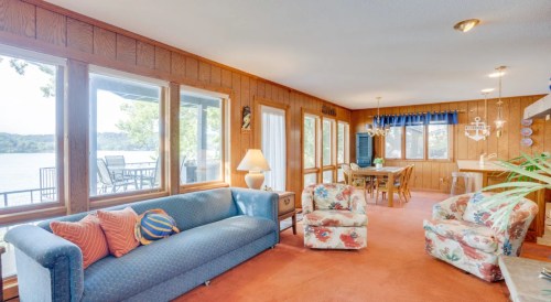 Cozy living area with a blue sofa, floral chairs, and large windows overlooking a lake. Warm wood paneling and bright decor.