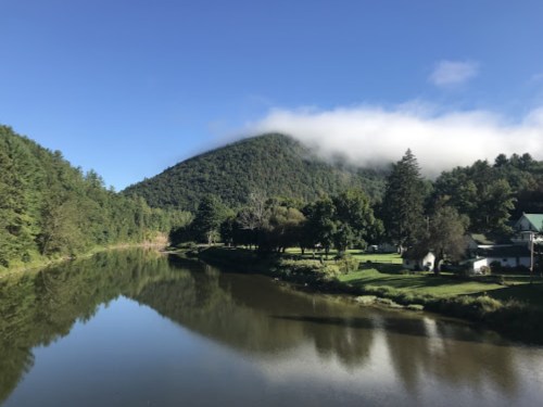 A serene river reflects a lush green mountain with a misty peak under a clear blue sky.
