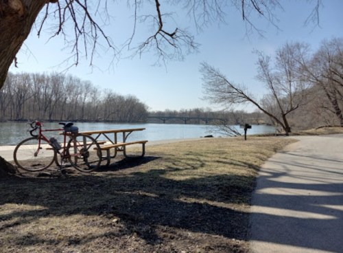 A peaceful riverside scene with a bike parked near a picnic table, trees lining the water's edge on a sunny day.