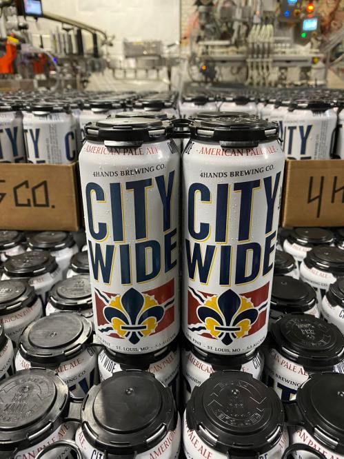 Cans of "City Wide" American Pale Ale from 4 Hands Brewing Co. stacked in a brewery setting.