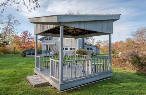 A wooden gazebo with a roof and chairs, surrounded by green grass and autumn trees in the background.
