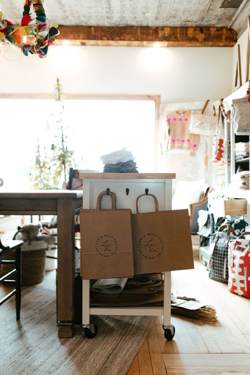 A cozy shop interior featuring a cart with paper bags, shelves of fabric, and natural light streaming through the window.