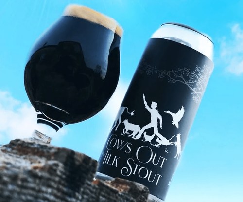 A glass of dark stout beer next to a can labeled "Cows Out Milk Stout," set against a blue sky.