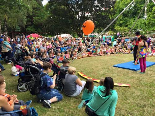 A large crowd gathers in a park for a performance, with children and families seated on the grass, enjoying the show.