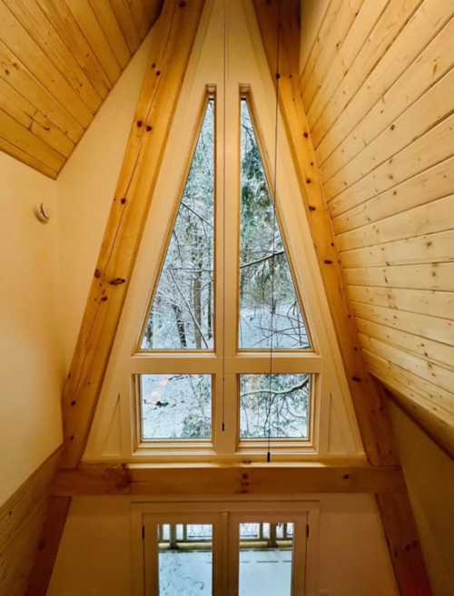 A triangular window in a wooden cabin, overlooking a snowy forest. Natural light fills the cozy interior.