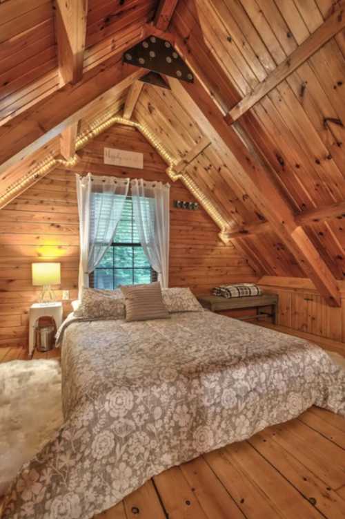 Cozy wooden bedroom with a patterned bedspread, large window, and warm lighting in a rustic cabin setting.