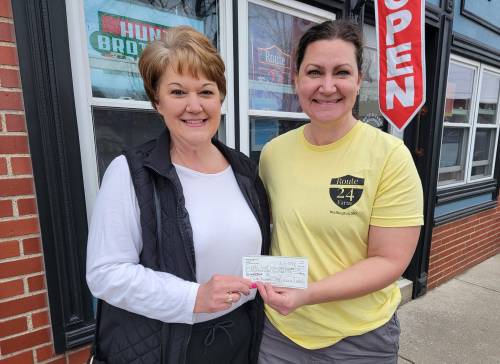 Two women stand outside a shop, smiling and holding a check together. A "Open" sign is visible in the background.