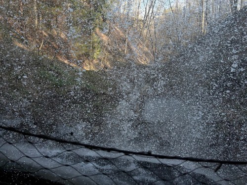 A view of a waterfall splashing against rocks, surrounded by trees and a sunny sky.