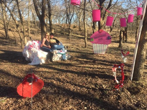 A couple sits on a decorated bench in a wooded area, surrounded by festive decorations and a large cupcake sign.
