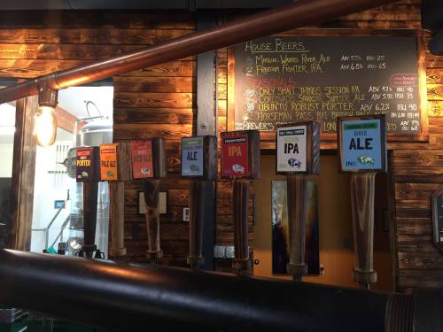 A wooden bar with various beer taps labeled with different beer types and prices displayed on a chalkboard behind.