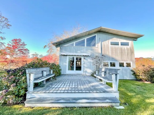 A modern, gray wooden house with large windows, surrounded by greenery and colorful autumn trees.