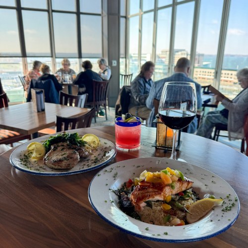 A table with two plates of seafood, a glass of red wine, and a colorful drink, with diners and a city view in the background.