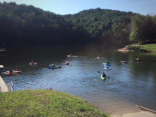 A serene lake scene with several kayakers paddling in the water, surrounded by green hills and trees.