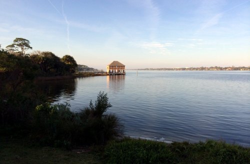A serene lake view with a wooden structure on stilts, surrounded by greenery and a clear blue sky.
