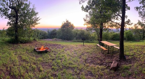 A serene campsite at sunset, featuring a fire pit, picnic table, and lush greenery with a distant view.