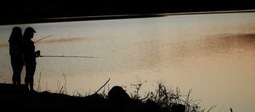 Two people fishing by a calm lake at sunset, silhouetted against the water's reflective surface.