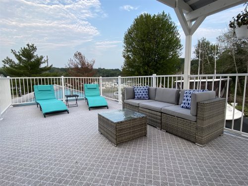 A cozy outdoor deck with a gray sofa, turquoise lounge chairs, and a glass coffee table, surrounded by trees.