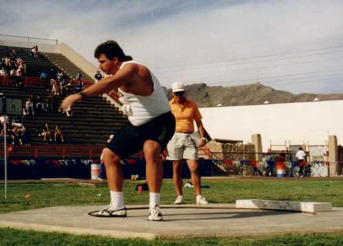 A male athlete prepares to throw a shot put, with a coach observing in a stadium setting.