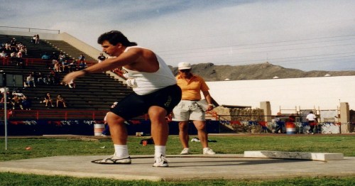 A male athlete prepares to throw a discus while a coach observes in a stadium setting.