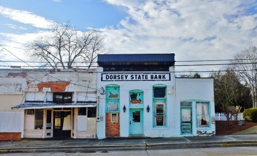 Historic Dorsey State Bank building with turquoise doors, surrounded by empty storefronts and a cloudy sky.