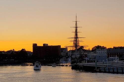 A tall ship silhouetted against a vibrant sunset over a calm harbor, with buildings in the background.