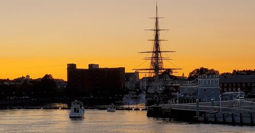 A sunset over a harbor with a tall ship and small boats, silhouetted against a vibrant orange sky.