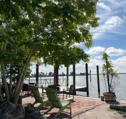 A serene waterfront scene with green chairs, trees, and a dock under a blue sky with fluffy clouds.