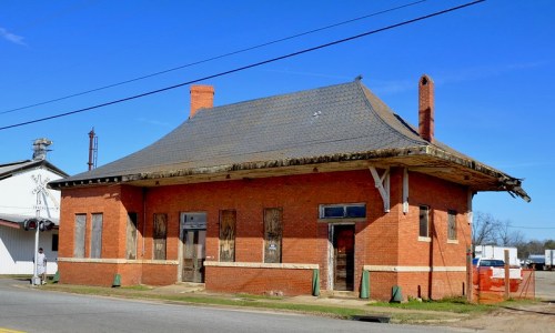 Abandoned red brick building with boarded windows and a sloped roof, set against a clear blue sky.
