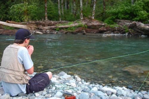 A person sits on a rocky riverbank, fishing with a rod, surrounded by trees and a flowing river.