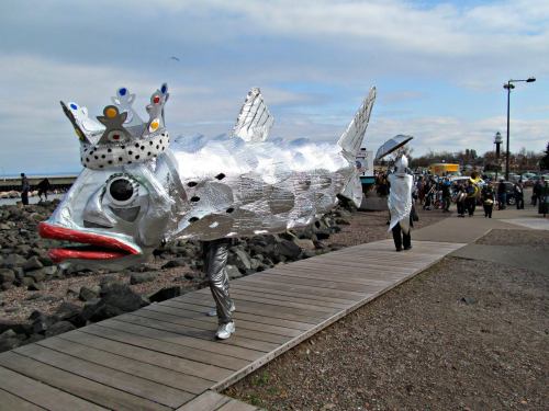 A person in a silver fish costume with a crown walks along a boardwalk, accompanied by others in the background.