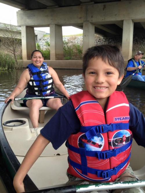 A boy smiles in a canoe with a woman behind him, both wearing life jackets, on a calm waterway under a bridge.